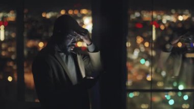 Medium arc shot of Afro-American businessman in suit and glasses standing by window in dark office at midnight, rubbing forehead and using smartphone, with bright bokeh cityscape in background