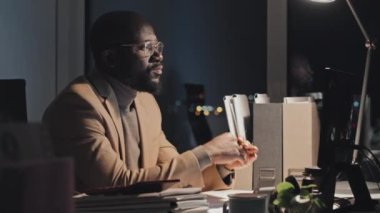 Medium shot of tired black male manager in beige suit jacket sitting at desk alone late at night in dark office, working on computer, then taking off glasses and rubbing eyes