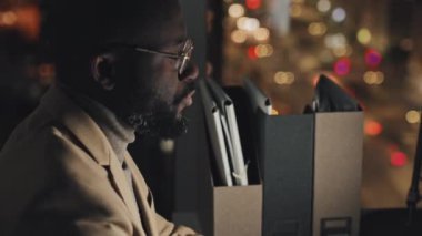 Medium close-up tiltnig down shot of face and hands of African American man in suit and glasses sitting at desk at night in city office and typing on computer keyboard