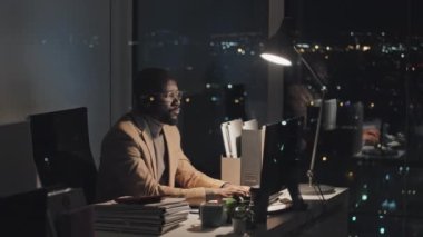 Full shot of sleepy Afro-American office worker sitting at desk writing document on computer, then taking off glasses and rubbing his eyes and face, with nighttime cityscape behind window