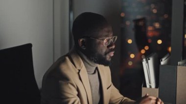 Chest-up shot of middle-aged black man sitting at desk, typing on computer, reading, then taking off glasses and rubbing face and eyes by hand, with bokeh nighttime city lights in background