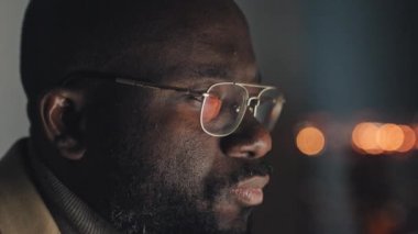 Close-up shot of face of exhausted bald African American man with beard and moustache looking at computer, then taking off glasses and rubbing his eyes, with bokeh lights in dark background