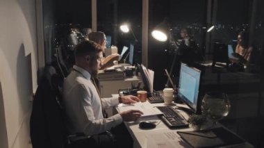 Full side shot of office workers sitting at desks, catching deadlines at night in office, Caucasian man looking through documents and loosening tie, and Muslim woman in hijab working on computer