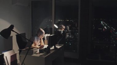 Müslüman tesettürlü kadın ofis çalışanının akşam geç saatlerde pencere kenarında tek başına oturması, bilgisayardaki belgeleri okuması ve evrak okuması, arka planda karanlık bir gece manzarası olması.