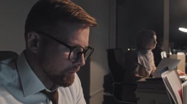 Close-up shot of face of young Caucasian male employee looking at computer screen late in evening in dark office, then massaging his temples, taking off glasses and blinking with strained eyes