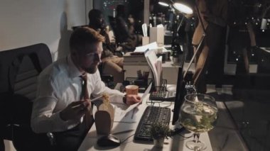 Medium shot of male corporate employee sitting at desk, reading document and working on computer, then taking mouthful of noodles out of takeaway box, with colleagues eating in background