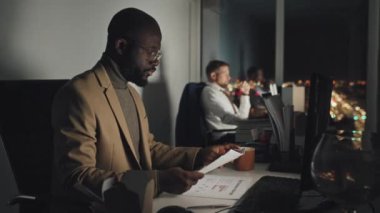 Medium shot of African American male project manager in suit and glasses sitting at desk in office at night and looking through freshly printed pages of presentation, then typing on computer