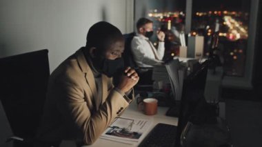 Medium shot of Afro-American male employee in suit and black protective facemask sitting at desk at night in office, reading document on screen, then putting on glasses and typing on keyboard