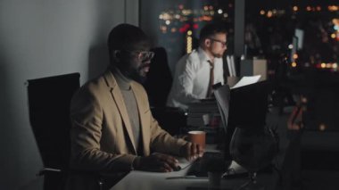 Medium shot of two multiethnic male employees sitting at desks in dark office and working on computers, then Afro-American man in glasses turning and looking at camera