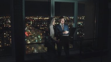 Full shot of Arab woman in Islamic hijab and Caucasian man in suit and tie standing by office window late in evening with laptop computer, talking, pointing at screen, smiling and laughing