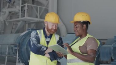 Medium shot of joyful Caucasian man and African American woman working together in marble factory watching something funny on digital tablet