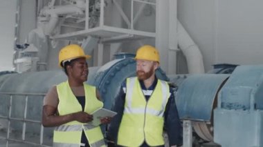 Slow motion of modern Caucasian man and Black woman in uniforms and hardhats walking along factory workshop discussing plans for work
