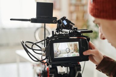 Screen of camera and hand of cameraman standing in spacious living room with comfortable couch, table and shelves with books