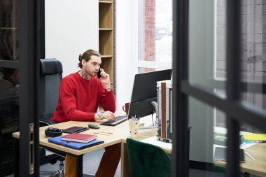 Serious young manager having a conversation on mobile phone while working on computer at his workplace at office