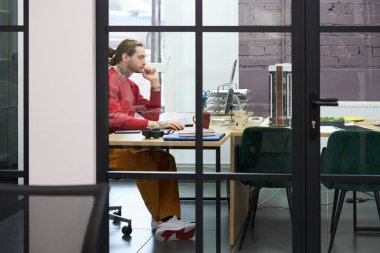 Young manager sitting at desk and working on computer behind the glass wall at modern office