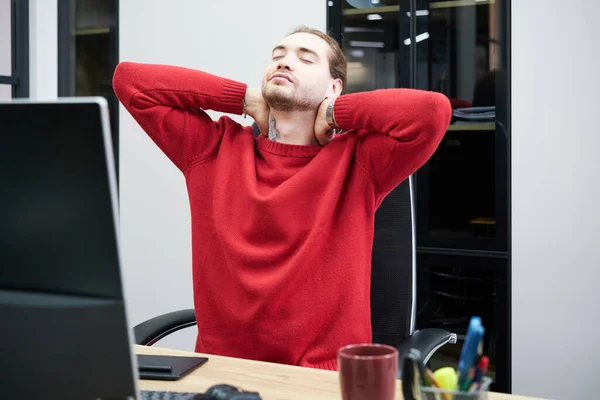 Young man stretching at his workplace in front of computer, he tired of work day at office
