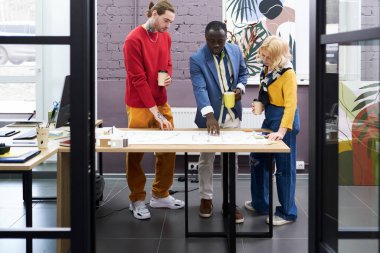 Group of professional architects examining new project on paper on table and discussing it during teamwork at office