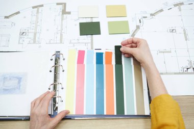 Close-up of interior designer choosing colour from colour palette while sitting at table with blueprint of new apartments