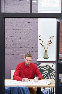 Young architect using ruler to draw blueprint of new construction project while sitting at workplace at office