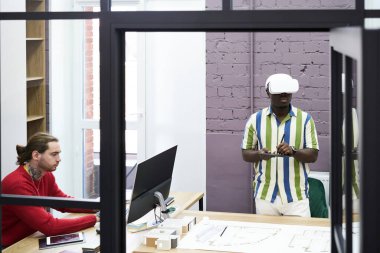 Graphic designer in vr headset working on tablet with his colleague working on computer at office study