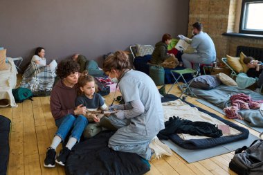 Healthcare worker in blue uniform, protective mask and gloves giving medical help to young female refugee and little boy in camp