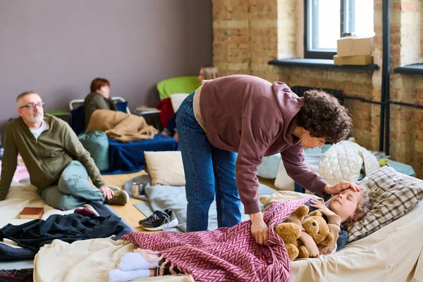 ホームレスの人々のために用意された広々とした部屋でソファの上で眠っている彼女の病気の息子を曲げ若い女性難民 — ストック写真