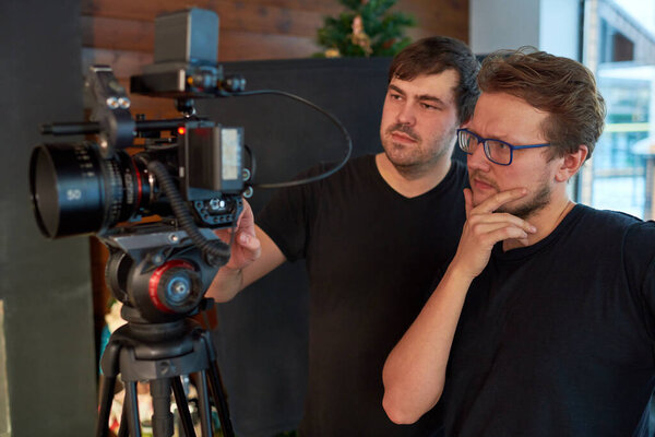 Young pensive videographer or operator and his assistant adjusting video camera while preparing for shooting of advertisement