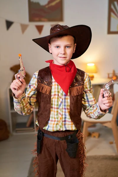 Happy cute little boy in costume of cowboy posing for commercial with two revolvers while standing in his room and looking at camera