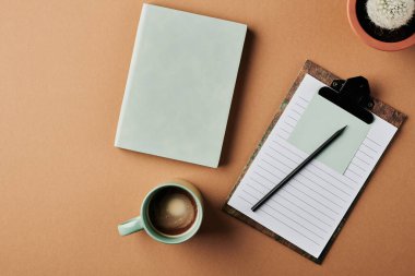 Top view of cup of coffee, clibboard with notepaper and document, small cactus in flowerpot and notebook over beige background