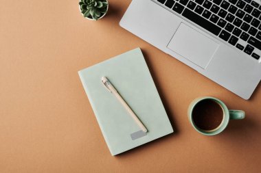 Overview of laptop keyboard, cup of coffee, notebook with pen and green plant in small flowerpot on workplace of modern business person