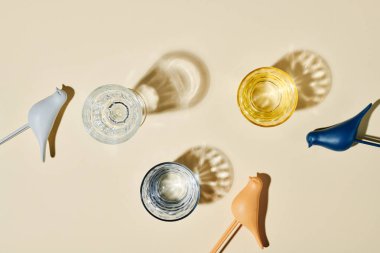 Flatlay of three glasses containing water, their shadows and small toy birds of various colors over beige background or on table
