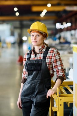 Young serious female worker of modern factory in hardhat and coveralls standing by part of huge industrial machine in workshop