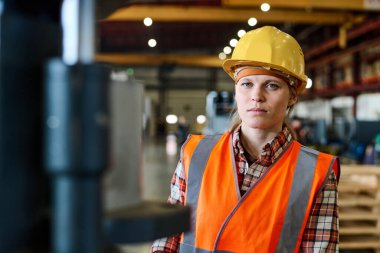 Young female worker of modern factory in orange jacket and protective helmet looking at camera in large workshop or warehouse