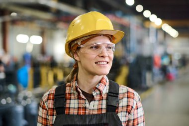 Happy young female engineer in hardhat and coveralls standing in workshop or warehouse of factory with industrial equipment