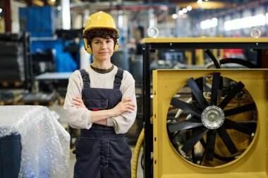 Young confident female engineer in workwear and protective headphones and helmet crossing arms by chest while standing in workshop
