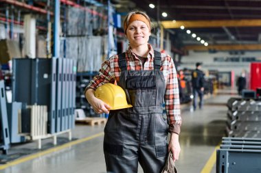 Happy young female engineer in coveralls and checkered shirt holding protective helmet in hand while standing in front of camera