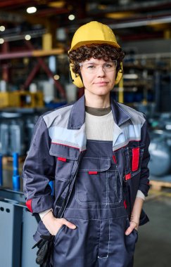 Young female worker of factory warehouse in uniform, protective helmet and headphones standing against new industrial equipment