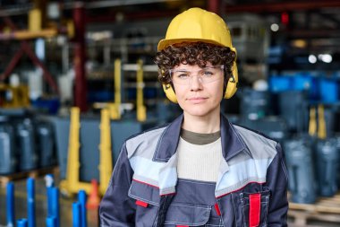 Young confident female engineer wearing uniform, hardhat, protective eyeglasses and headphones standing in front of camera