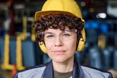 Face of young engineer in protective helmet, eyeglasses and headphones looking at camera while standing against industrial equipment
