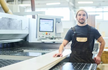 Portrait of a young carpenter in an apron, smiling at the camera, standing at the modern furniture production equipment