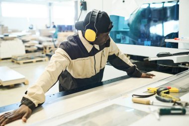 African carpenter in protective headphones and uniform sawing wooden plank on special machine at factory