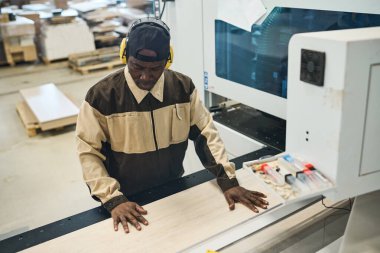 African craftsman in protective headphones and uniform concentrating on his wood work at machine