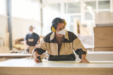 Female craftsperson in protective mask and headphones using tape measure to examine the length of wooden plank