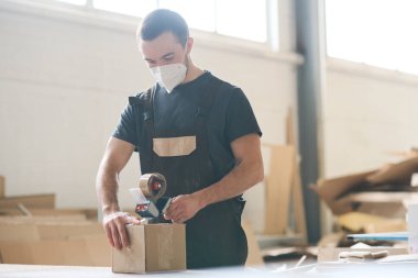 Young worker in protective mask working in distribution warehouse, he packing boxes with adhesive tape