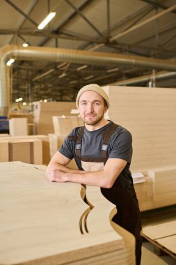 Portrait of young carpenter in overalls looking at camera standing with finished wooden products at furniture factory
