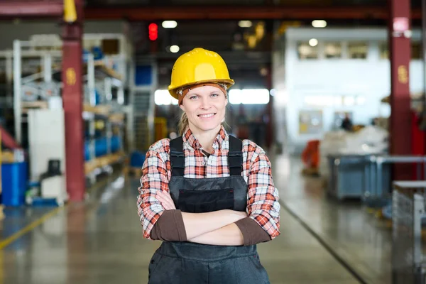 Happy young female engineer of modern factory in hardhat and coveralls crossing arms by chest while standing in front of camera