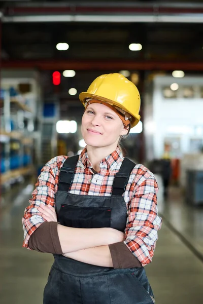 Young successful female engineer in workwear standing in spacious workshop of large modern factory with her arms crossed by chest