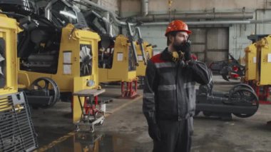 Medium long of Caucasian man in orange hard hat and uniform using walkie talkie, standing at tractor plant at daytime