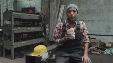 Medium long portrait of female Hispanic plant worker holding sandwich, sitting at factory in afternoon, looking on camera