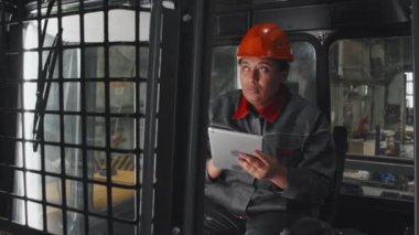 Medium long of African American woman in orange hard hat and work overalls sitting inside cabin of new machinery at factory, inspecting it, using tablet